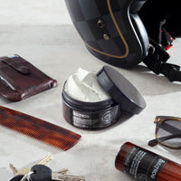 Bullfrog Strong Adhesion Matte Wax, boîte ouverte avec cire blanche en flat lay avec casque, peigne et lunettes
