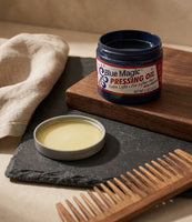 Blue Magic Pressing Oil - pot ouvert sur une ardoise avec un peigne en bois, l'huile jaune visible